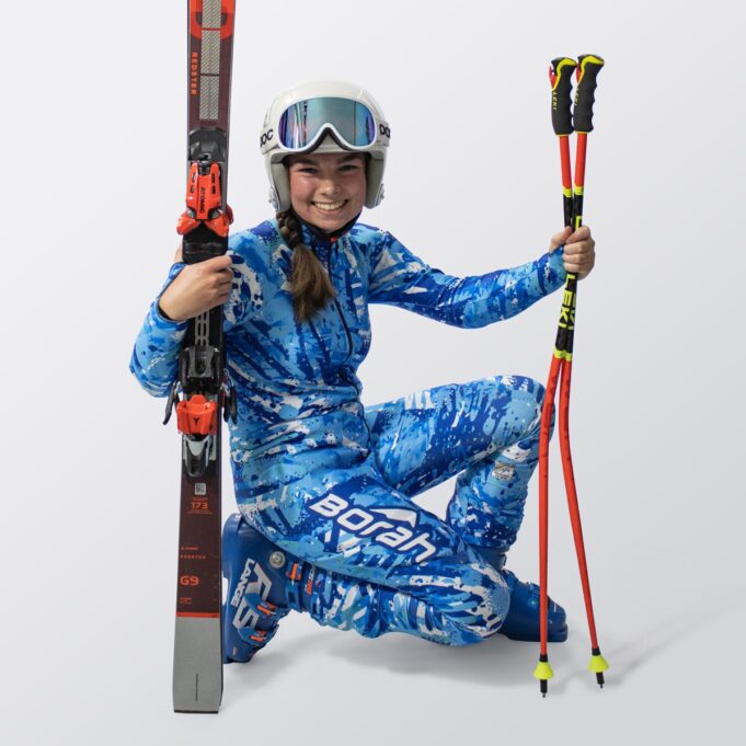 A young female athlete crouching while holding her skis and poles wearing a custom alpine race suit.