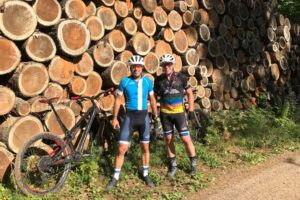 Two cyclists posing near a pile of timber.