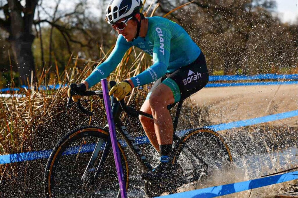Cyclocross athlete wearing a custom cycling kit, shredding through a large mud puddle, splashing water and muck up.