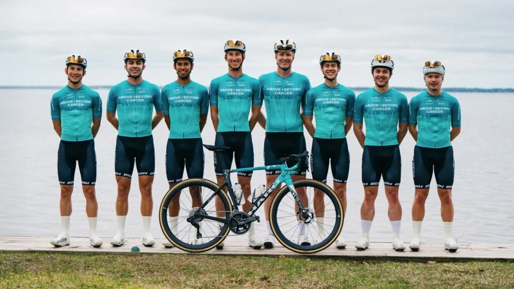 Group photo of the Above and Beyond Cancer cycling team posing in their new custom cycling jerseys with a bike in front of a lake.