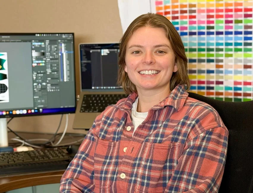 a female wearing a plaid shirt smiles and sits in front of a computer workstation. A wall of colors is in the background.