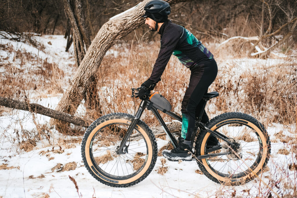 Winter cyclist riding through the snow on a black fat bike wearing black Custom Borah Teamwear apparel. Snow and woodlands in the background.