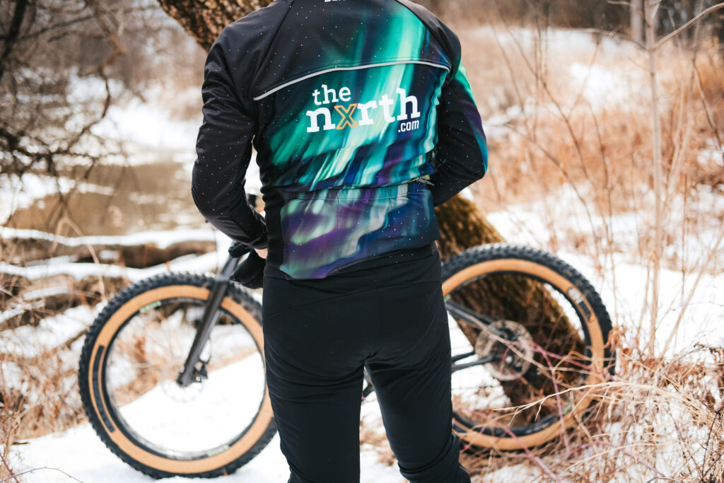 A person facing away from the camera, wearing a custom OTW Thermal Cycling Jacket by Borah Teamwear. A fatbike leans against a tree in the background.