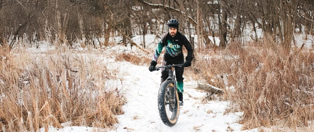 A wide shot of a cyclist wearing black Borah gear, and riding a black fat bike, riding on a snowy trail through the woods.
